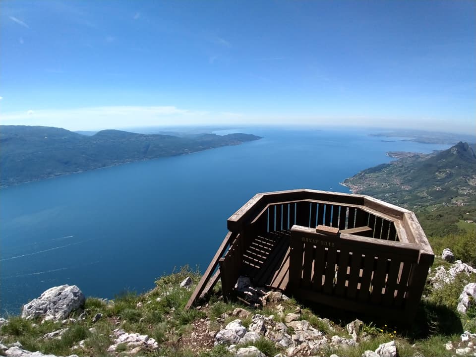 Wooden panoramic balcony on Cima Comer offering a spectacular view over Lake Garda on a sunny day.
