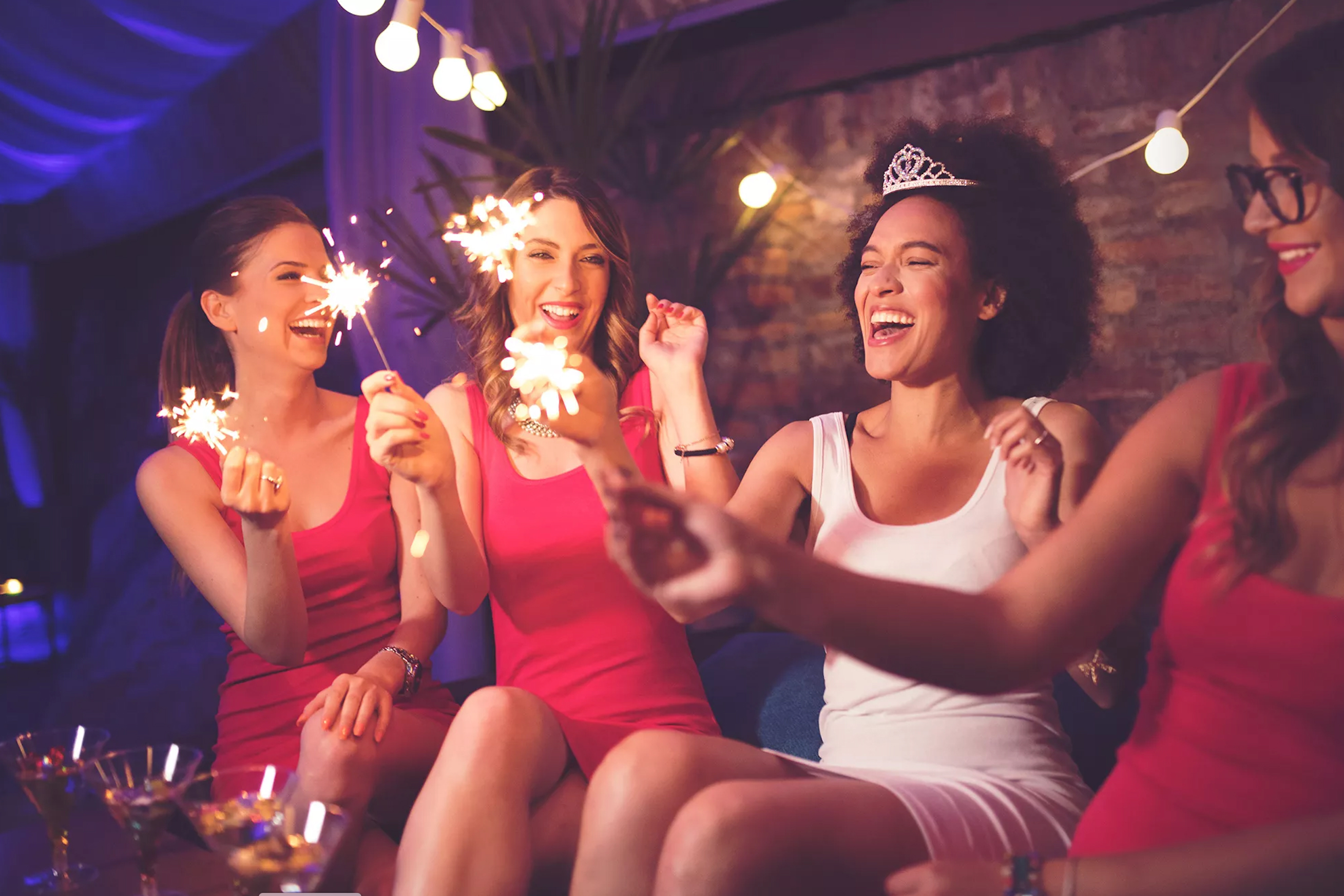 Smiling women celebrating a bachelorette party with sparklers and drinks in a dimly lit venue.