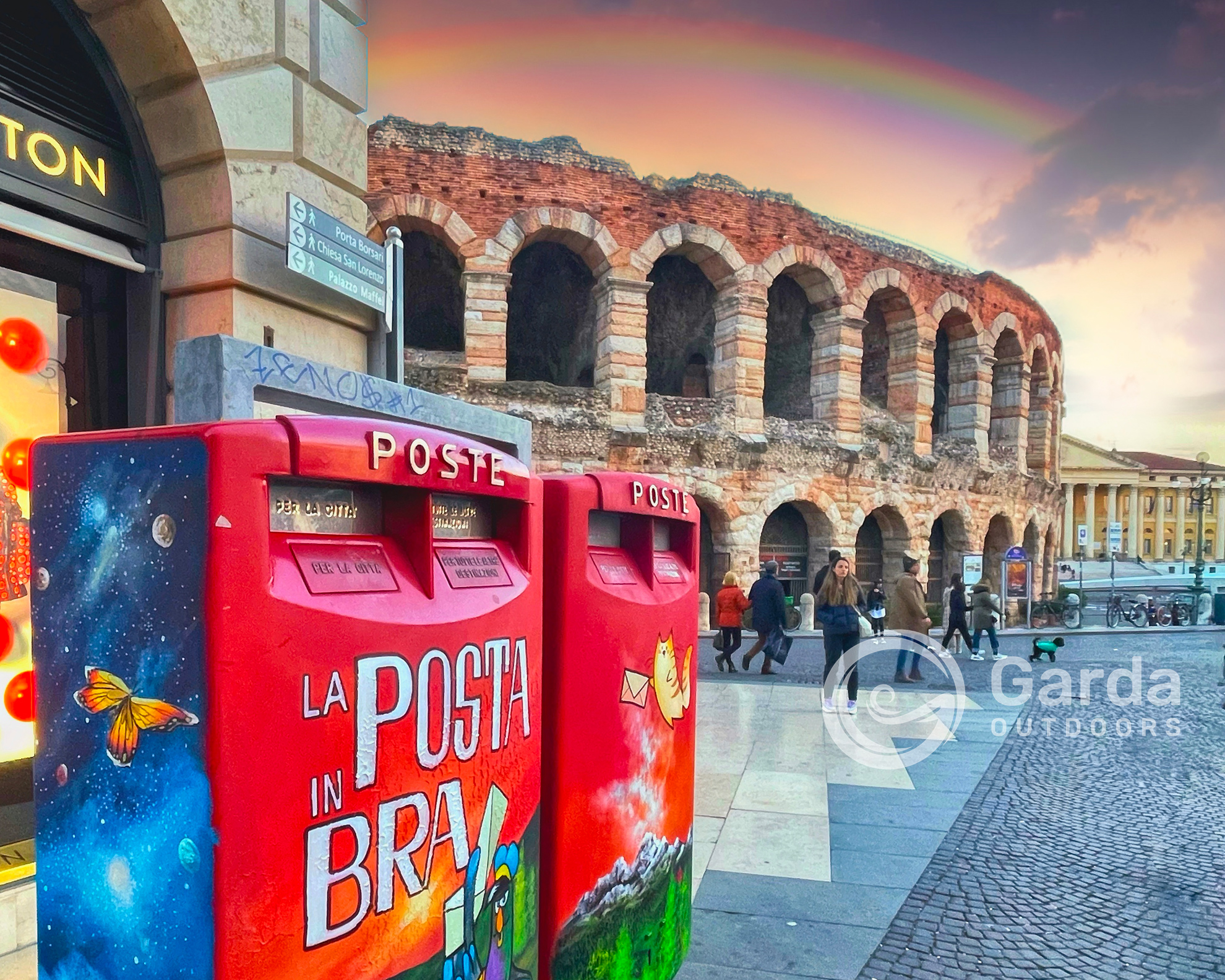 Red decorated mailboxes in Piazza Bra with the Verona Arena and a rainbow in the background.